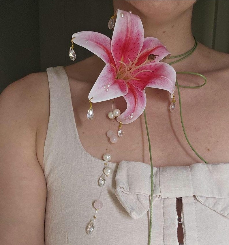 May include: A large pink and white lily flower brooch featuring dangling clear crystal teardrops and round white pearls. The floral accessory is attached to a thin green cord that wraps around the neck, accented with additional pearls and crystals.