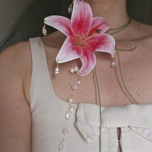 May include: A large pink and white lily flower brooch featuring dangling clear crystal teardrops and white pearls. The floral piece is attached to a thin green cord that wraps around the neck, accented with a vertical strand of pearls and crystals.