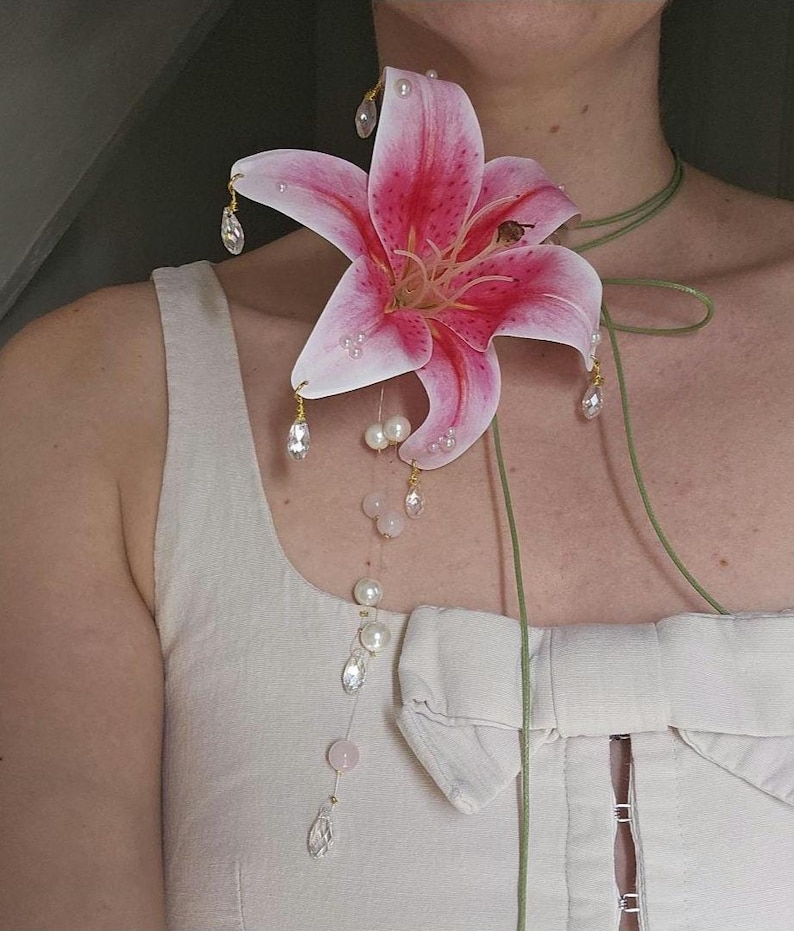 May include: A large pink and white lily flower brooch featuring dangling clear crystal teardrops and white pearls. The floral piece is attached to a thin green cord that wraps around the neck, accented with a vertical strand of pearls and crystals.