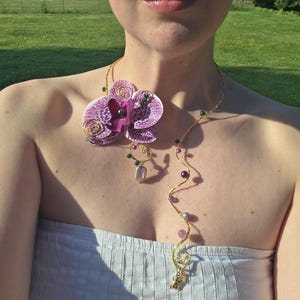 Op de afbeelding: Een goudkleurige ketting met een grote, paarse orchideehanger met groene en paarse accenten. De ketting heeft een delicaat, kronkelend ontwerp met paarse en groene kralen en een kleine bedel aan het einde. De ketting wordt gedragen over een lichtblauwe strapless top.