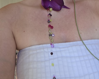 May include: A choker necklace featuring a deep purple calla lily flower attached to a thin green cord. A long, vertical strand of multicolored beads in shades of pink, purple, yellow, and dark grey hangs downward from the center of the flower.