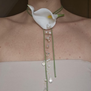 Puede incluir: Una flor de lirio blanco con tallo y hojas verdes está unida a un collar de cordón verde con cuentas blancas y transparentes. El collar lo lleva una persona.