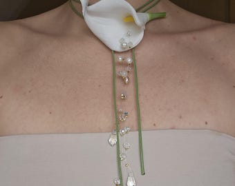 May include: A choker necklace featuring a white calla lily flower with a yellow spadix, attached to thin green cords. A vertical strand of clear crystal beads and small white pearls hangs downward from the center of the flower, creating a long pendant.
