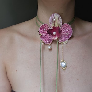 Puede incluir: Un collar único con forma de flor de orquídea. Presenta una gran flor morada y blanca, un cordón verde, pequeñas estrellas doradas, perlas colgantes y un colgante de hoja dorada con una perla grande.