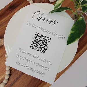 May include: A white card with black text that reads "Cheers To the Happy Couple" and a black and white QR code. The text below the QR code reads "Scan the QR code to buy them a drink on their Honeymoon".