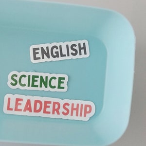 May include: A blue tray with three white stickers on it. The stickers say "ENGLISH", "SCIENCE", and "LEADERSHIP".