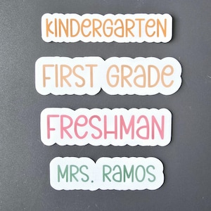 May include: Four white stickers with rounded corners and black outlines. The stickers have different colored text. The top sticker says "Kindergarten" in orange. The second sticker says "First Grade" in light orange. The third sticker says "Freshman" in pink. The bottom sticker says "Mrs. Ramos" in green.