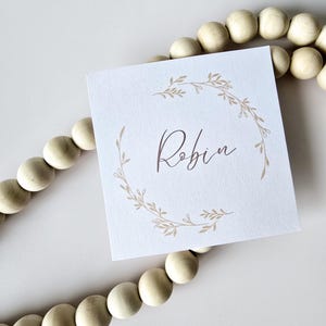 May include: A white card with a brown floral wreath and the name "Robin" written in brown cursive.