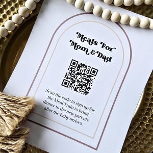 Peut inclure: Une affiche blanche imprimée avec le texte "Meals For Mom & Dad" et un code QR. L'affiche est posée sur un plateau doré, avec une guirlande de perles en bois et un paquet de ficelle en arrière-plan. Le texte de l'affiche encourage les gens à s'inscrire à un repas.