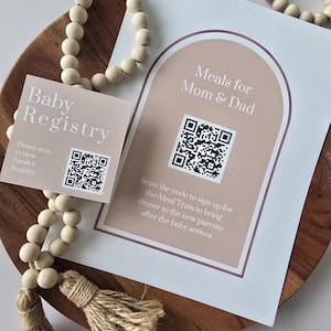 Pode incluir: Uma grinalda de contas de madeira e dois cartões numa bandeja de madeira. Um cartão diz "Baby Registry" e o outro "Meals for Mom & Dad". Ambos os cartões têm códigos QR. O texto no segundo cartão incentiva a digitalizar o código para se inscrever num comboio de refeições.