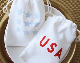 Könnte beinhalten: Zwei weiße Kordelzugbeutel. Einer zeigt eine Ansammlung hellblauer Sterne, der andere die roten Buchstaben "USA". Beide Beutel liegen auf einem weißen Teller vor einem goldfarbenen, strukturierten Hintergrund.
