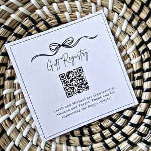 May include: A white card with a black bow and the text "Gift Registry" printed on it. A black and white QR code is below the text. The text below the QR code reads "Sarah and Michael are registered at Amazon and Target. Thank you for supporting the happy couple!"