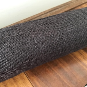 May include: A rolled-up, dark grey fabric with a woven texture. The fabric rests on a wooden surface, with a glimpse of a white wall in the background. The close-up shot highlights the detailed weave of the material.