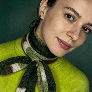 May include: A person wearing a lime green sweater and a striped scarf with shades of green, brown, and white. The scarf is tied in a knot. The background is a dark green.