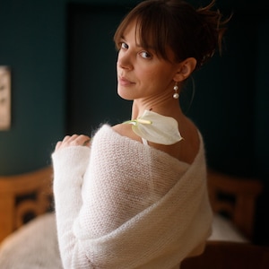 May include: A woman wearing a white, fluffy knit wrap. A white calla lily rests on her shoulder. She is wearing pearl earrings and a white dress. The background is a dark teal color.