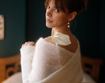 May include: A woman wearing a white, fluffy knit wrap. A white calla lily rests on her shoulder. She is wearing pearl earrings and a white dress. The background is a dark teal color.
