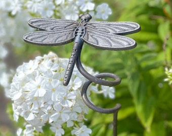 Puede incluir: Una estaca de jardín de metal en forma de libélula con un tallo rizado. La libélula está pintada de plata y tiene detalles negros. La estaca está rodeada de flores blancas.