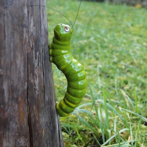 Könnte beinhalten: Eine grüne Keramikraupe mit schwarzen Augen und einem gelben Streifen, die an einem Holzpfosten hochklettert. Die Raupe befindet sich in einem grasbewachsenen Bereich.