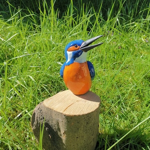 Könnte beinhalten: Eine Keramik-Vogelfigur, die in Blautönen, Orange und Weiß bemalt ist, sitzt auf einem Holzstumpf. Der Vogel hat einen langen, dünnen Schnabel und ist mit dem Kopf nach oben geneigt.