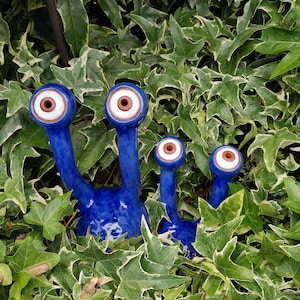 Könnte beinhalten: Drei blaue Keramik-Gartenornamente mit großen weißen Augen und braunen Pupillen, die aus einem Bett aus grünen Blättern ragen.