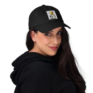 Peut inclure: Une casquette de baseball noire avec un écusson carré blanc sur le devant. L'écusson présente un graphique de soleil jaune, une silhouette de palmier noir et le texte EJ7S en lettres noires grasses. La casquette a une visière courbée.