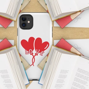 Puede incluir: Funda de teléfono blanca con dos globos rojos en forma de corazón y la palabra "LOVE". Rodeada de libros abiertos y lápices rojos sobre una superficie marrón claro.