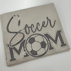 Peut inclure: Un t-shirt beige avec les mots "Soccer MOM" en strass noirs. Le mot "Soccer" est écrit en cursive, et un ballon de football est au centre du mot "MOM".