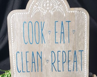 Könnte beinhalten: Ein Holzschild mit einer abgenutzten weißen Farbe und einer blauen Aufschrift, die "Cook Eat Clean Repeat" lautet. Das Schild hat einen Ständer und ist oben mit einem weißen Blumenmuster verziert. Im Vordergrund befinden sich eine Reihe von Holzperlen und ein Jute-Quaste.