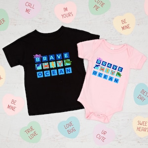 Op de afbeelding: Een zwart T-shirt en een roze bodysuit met de woorden "Brave Ocean" in een blokontwerp. Het ontwerp bevat een verscheidenheid aan zeedieren, waaronder een krab, een dolfijn, een schildpad en een zeester.