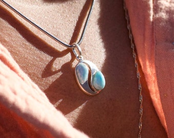 May include: A silver necklace with a pendant featuring two light blue and white teardrop-shaped stones set in a gold-colored metal. The necklace is worn over a peach-colored shirt.