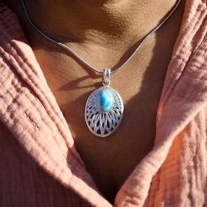 May include: A silver pendant necklace featuring an oval-shaped, light blue gemstone. The pendant is set in a decorative, openwork silver frame. The necklace is displayed against a peach-colored fabric.