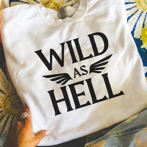 以下が含まれることがあります： 「WILD AS HELL」の文字が黒で大胆にプリントされた白いスウェットシャツ。翼のイラストが添えられています。模様のある表面に折りたたまれており、手と指輪が見えます。