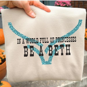 Peut inclure: Un sweat-shirt beige avec le texte "IN A WORLD FULL OF PRINCESSES BE A BETH" en noir et un motif de cornes de taureau en paillettes turquoise. Des citrouilles et des fleurs en pot sont en arrière-plan.