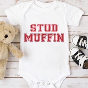 May include: White baby onesie with red text that reads "Stud Muffin" in a collegiate font.