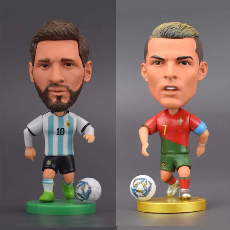 Może przedstawiać: Dwie figurki bobblehead znanych piłkarzy, Lionela Messiego i Cristiano Ronaldo. Messi ma na sobie jasnoniebieską i białą koszulkę reprezentacji Argentyny z numerem 10. Ronaldo ma na sobie czerwoną i zieloną koszulkę reprezentacji Portugalii z numerem 7. Obie figurki trzymają piłkę nożną.