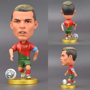 Może przedstawiać: Figurka bobblehead piłkarza w czerwono-zielonej koszulce z numerem 7 z przodu. Piłkarz trzyma piłkę nożną i stoi na złotej podstawie.