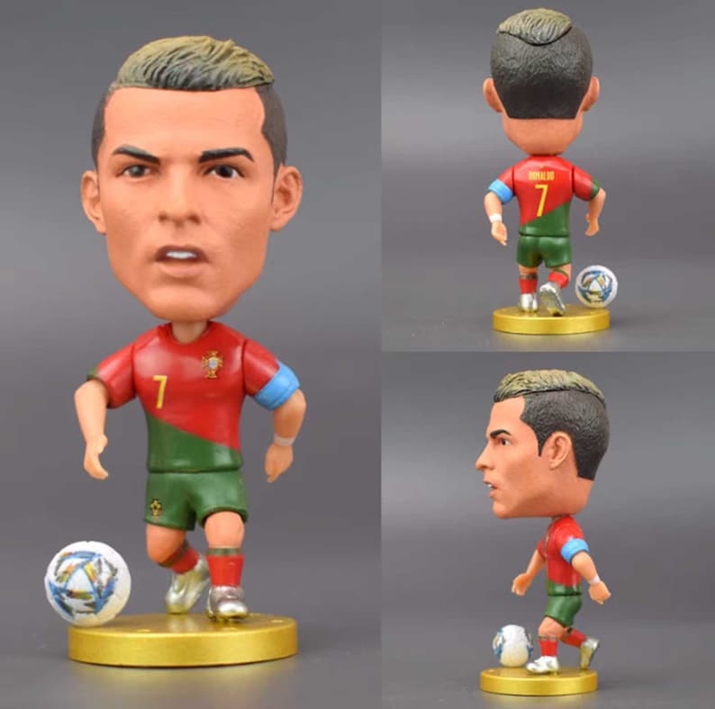 Może przedstawiać: Figurka bobblehead piłkarza w czerwono-zielonej koszulce z numerem 7 z przodu. Piłkarz trzyma piłkę nożną i stoi na złotej podstawie.