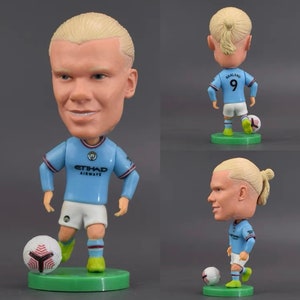 Może przedstawiać: Niebieska i biała figurka bobblehead piłkarza z blond warkoczem. Figurka ma na sobie niebieską koszulkę z napisem "ETIHAD AIRWAYS" i numerem "9" z tyłu. Figurka trzyma piłkę nożną i stoi na zielonej podstawie.