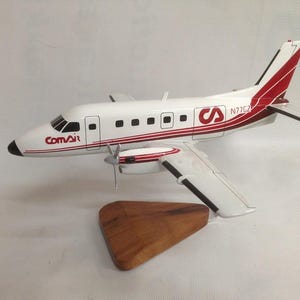 May include: A white and red Comair airplane model with a wooden base. The model features the Comair logo and the registration number N77CZ. The airplane has a propeller and a red stripe along the fuselage.