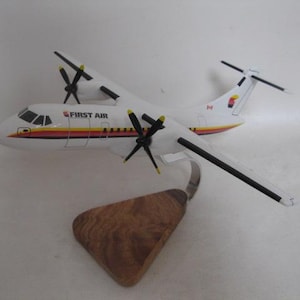 May include: A model airplane of a First Air ATR 42-300, a twin-turboprop regional airliner. The model is white with black, yellow, and red stripes. The plane has four propeller blades and is mounted on a wooden stand.
