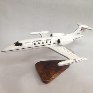 May include: A white model airplane with the text "UNITED STATES OF AMERICA" on the side. The model is mounted on a wooden base. The plane has a sleek design with wings and tail fins.