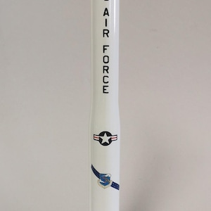 May include: A white model rocket with the text "US AIR FORCE" printed vertically. The rocket features a star emblem and a blue and white insignia. It stands on a black circular base.