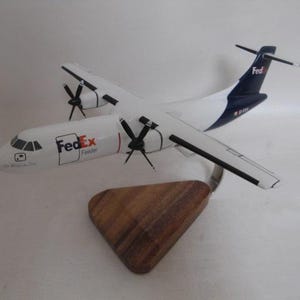 May include: A white and blue FedEx Feeder airplane model with black propellers and a wooden stand. The model has the FedEx logo on the side and is a detailed replica of a cargo aircraft.