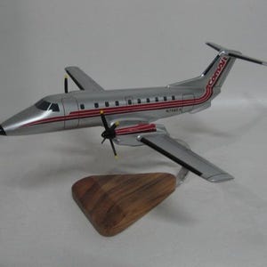 May include: A silver and red model airplane with the word "Comair" on the tail. The model has two propellers and is mounted on a wooden stand. The airplane has red stripes and windows.