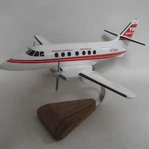 May include: A white and red model airplane with the text "TRANS WORLD EXPRESS" and "N71999" printed on the fuselage. The model is mounted on a wooden base and has a silver support.