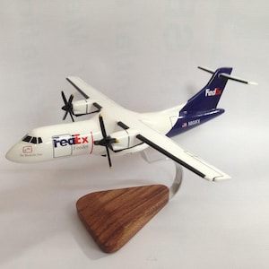 May include: A white and blue FedEx Feeder airplane model with a wooden base. The model has two propellers and the FedEx logo on the side and tail. The text "The World On Time" is on the nose.