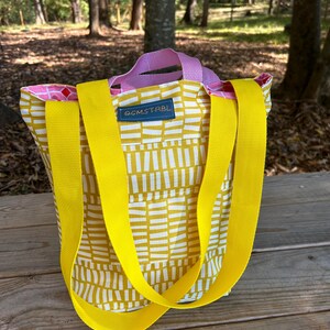 Könnte beinhalten: Eine gelbe und weiße gemusterte Tragetasche mit gelben Trägern und einem rosafarbenen Griff. Die Tasche hat ein kleines blaues Etikett mit der Aufschrift "COMSTRBL".