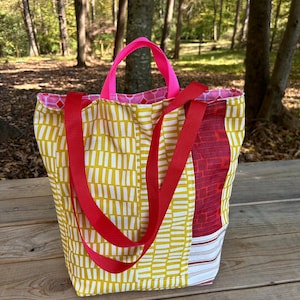 Könnte beinhalten: Eine gelbe und rote Tragetasche mit pinkfarbenen Henkeln. Die Tasche hat ein geometrisches Muster und ist aus Stoff.