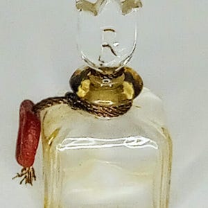 May include: A small, square, clear glass bottle with a light yellow tint. The bottle features a clear glass stopper shaped like a tulip. A thin, dark brown cord is wrapped around the neck, secured with a small, rectangular red wax seal.