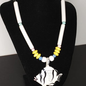 May include: A white ceramic necklace with a black and white fish pendant. The necklace is made of white, yellow, pink, and blue beads.
